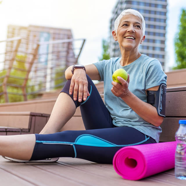 Woman smiles while holding an apple while sitting next to a yoga mat. She is eating healthy and exercising.